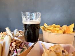 A tall glass filled with a dark beverage, possibly stout, sits on a table alongside a bowl of crispy potato chips. Nearby, there are serving trays containing neatly arranged bao buns filled with shredded meat and vegetables.