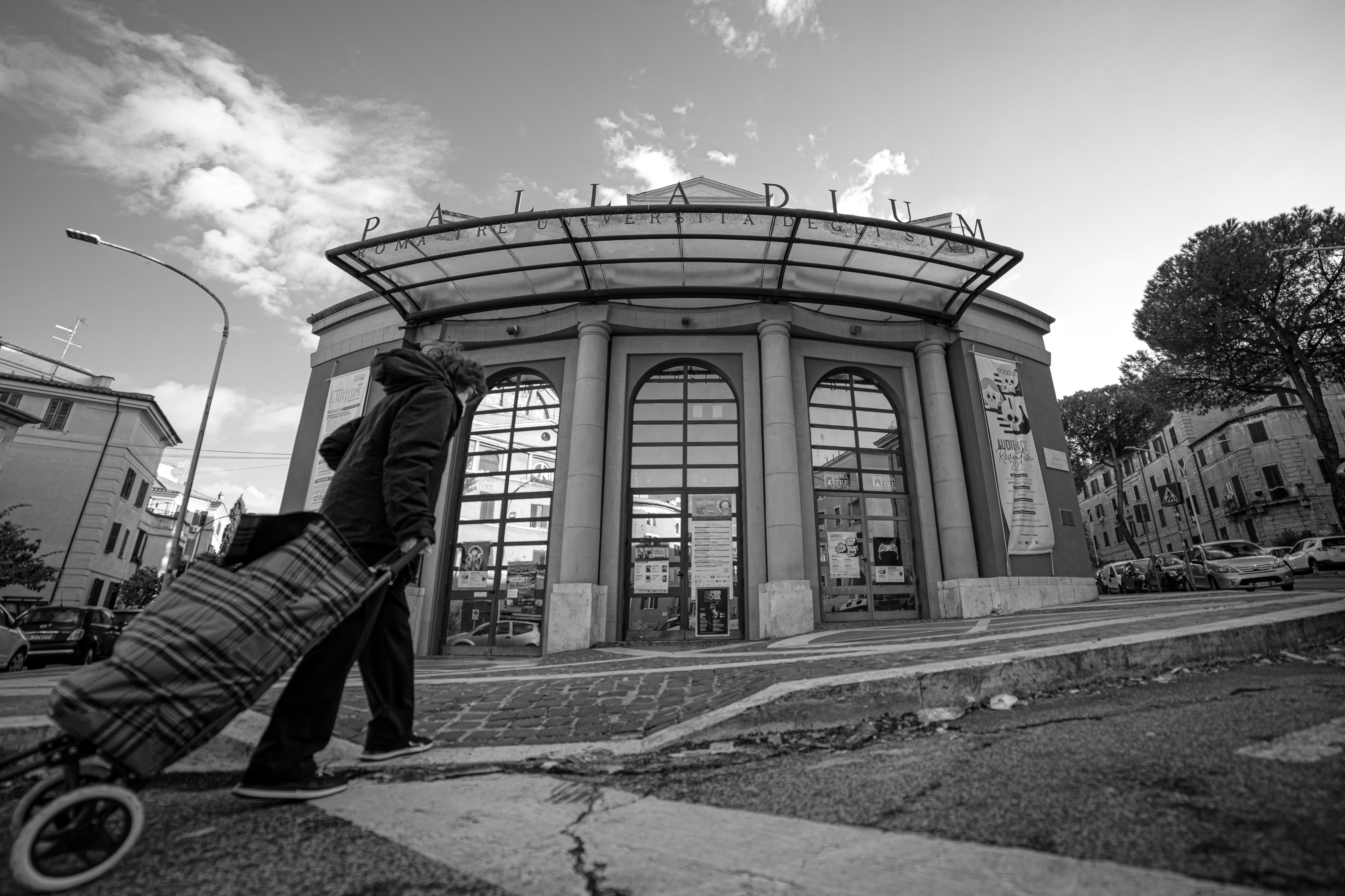a person pushing a cart, A wide angle photo in black and white of a lady with a cart passing in front of a theater in Rome.