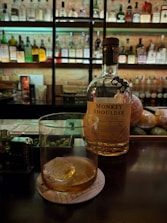 A dimly lit bar with a bottle of Monkey Shoulder whiskey placed on the counter next to a glass containing a drink with ice. Shelves in the background are filled with an array of various liquor bottles. Limes and oranges are seen on the counter near the bottle.