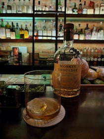 A dimly lit bar with a bottle of Monkey Shoulder whiskey placed on the counter next to a glass containing a drink with ice. Shelves in the background are filled with an array of various liquor bottles. Limes and oranges are seen on the counter near the bottle.