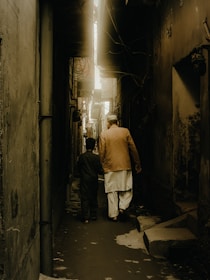 A narrow alley with high, dimly lit walls on either side. Two people, one older and one younger, walk side by side, creating a sense of companionship. The scene is illuminated by a soft, golden light that filters down from above, casting shadows along the path.