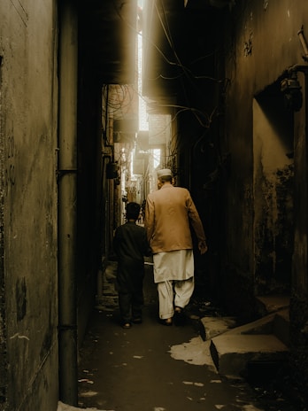 A narrow alley with high, dimly lit walls on either side. Two people, one older and one younger, walk side by side, creating a sense of companionship. The scene is illuminated by a soft, golden light that filters down from above, casting shadows along the path.