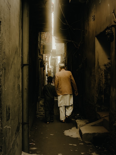 A narrow alley with high, dimly lit walls on either side. Two people, one older and one younger, walk side by side, creating a sense of companionship. The scene is illuminated by a soft, golden light that filters down from above, casting shadows along the path.
