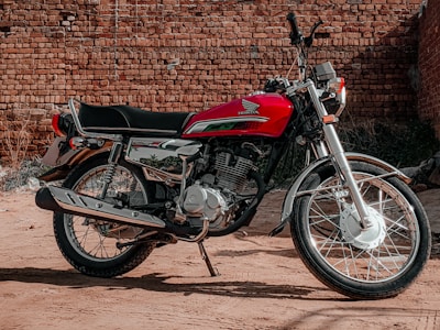 A red Honda motorcycle is parked on a dirt surface in front of a rustic brick wall. The motorcycle features chrome detailing and a sleek, vintage design with wire-spoked wheels.