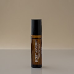 A brown glass bottle with a black cap stands vertically on a reflective surface. The label on the bottle reads 'SECRET ALCHEMIST' in white letters, along with smaller text beneath it. The background is a plain, neutral beige color.