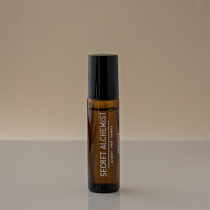 A brown glass bottle with a black cap stands vertically on a reflective surface. The label on the bottle reads 'SECRET ALCHEMIST' in white letters, along with smaller text beneath it. The background is a plain, neutral beige color.