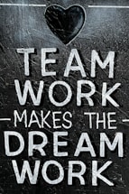 The image features the motivational phrase 'Team Work Makes The Dream Work' written in bold, white capital letters on a textured, dark background. A heart shape is positioned above the text, adding a positive and encouraging element.