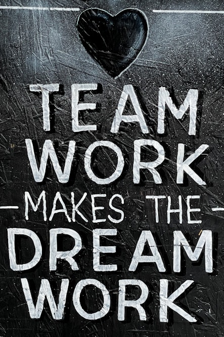 The image features the motivational phrase 'Team Work Makes The Dream Work' written in bold, white capital letters on a textured, dark background. A heart shape is positioned above the text, adding a positive and encouraging element.