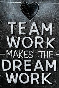 The image features the motivational phrase 'Team Work Makes The Dream Work' written in bold, white capital letters on a textured, dark background. A heart shape is positioned above the text, adding a positive and encouraging element.