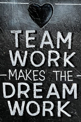 The image features the motivational phrase 'Team Work Makes The Dream Work' written in bold, white capital letters on a textured, dark background. A heart shape is positioned above the text, adding a positive and encouraging element.