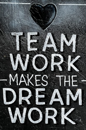 The image features the motivational phrase 'Team Work Makes The Dream Work' written in bold, white capital letters on a textured, dark background. A heart shape is positioned above the text, adding a positive and encouraging element.