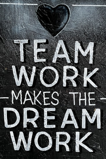 The image features the motivational phrase 'Team Work Makes The Dream Work' written in bold, white capital letters on a textured, dark background. A heart shape is positioned above the text, adding a positive and encouraging element.
