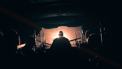 A group of silhouetted people sits inside a dark vehicle, possibly a military boat or aircraft, facing a bright, glowing light in the distance. The setting is moody, with shadows and dim lighting.