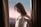 A woman in a white corset top stands between two brown curtains. She is looking downward with a subtle expression, holding one of the straps of her garment. The background is softly blurred in shades of pink and blue, suggesting a serene setting.