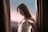 A woman in a white corset top stands between two brown curtains. She is looking downward with a subtle expression, holding one of the straps of her garment. The background is softly blurred in shades of pink and blue, suggesting a serene setting.