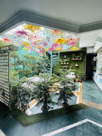 A vibrant indoor garden setup featuring numerous potted plants, a white table with chairs, and a spiral staircase painted light blue. The ceiling is adorned with colorful flower-shaped decorations in yellow, pink, orange, and blue. There are birdhouses and shelves with plants mounted on a green wall in the background.