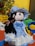 A plush doll with black yarn hair and a blue dress is sitting on a textured red surface. The doll has a matching blue hat and a small blue cat embroidered on its dress. Surrounding the doll are vibrant artificial flowers in pots, including yellow daisies and multicolored blossoms, contributing to a cheerful and playful setting.