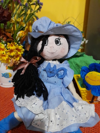 A plush doll with black yarn hair and a blue dress is sitting on a textured red surface. The doll has a matching blue hat and a small blue cat embroidered on its dress. Surrounding the doll are vibrant artificial flowers in pots, including yellow daisies and multicolored blossoms, contributing to a cheerful and playful setting.