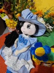 A handmade doll wearing a blue dress and hat, adorned with decorative flowers. It has long black hair and large embroidered eyes. Beside the doll is a plush blue flower with a smiling face. In the background, there are vibrant artificial flowers against a yellow backdrop, adding a cheerful and colorful feel.
