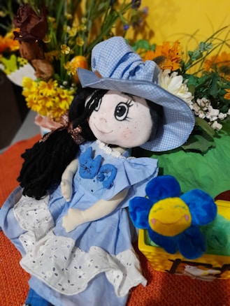 A handmade doll wearing a blue dress and hat, adorned with decorative flowers. It has long black hair and large embroidered eyes. Beside the doll is a plush blue flower with a smiling face. In the background, there are vibrant artificial flowers against a yellow backdrop, adding a cheerful and colorful feel.