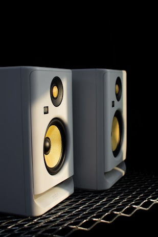 Two white speakers with yellow cones sit side by side on a metallic surface, illuminated by a soft light against a dark background. Both speakers have a compact design, featuring circular tweeters and woofers.