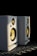 Two white speakers with yellow cones sit side by side on a metallic surface, illuminated by a soft light against a dark background. Both speakers have a compact design, featuring circular tweeters and woofers.