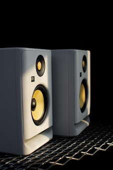 Two white speakers with yellow cones sit side by side on a metallic surface, illuminated by a soft light against a dark background. Both speakers have a compact design, featuring circular tweeters and woofers.
