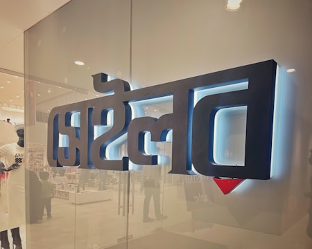 A glass storefront with large, black, three-dimensional text in a script style, backlit with white and blue lighting. The interior of a store is faintly visible through the glass, showcasing mannequins and various products.