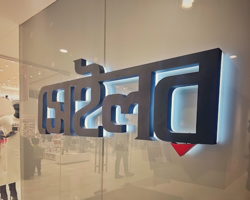 A glass storefront with large, black, three-dimensional text in a script style, backlit with white and blue lighting. The interior of a store is faintly visible through the glass, showcasing mannequins and various products.