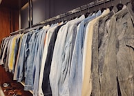 A neatly organized row of jackets hanging on a clothing rack, featuring a variety of denim, tan, and neutral colors. The wooden wall and shelves add a warm touch to the setting, with a few pairs of sandals placed below.