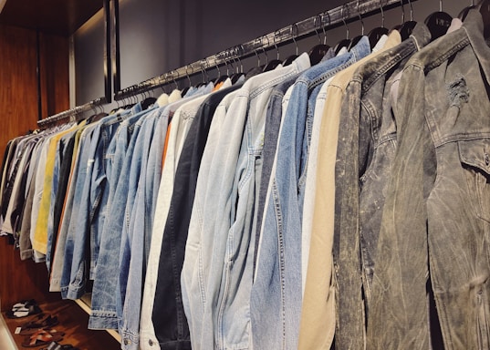 A neatly organized row of jackets hanging on a clothing rack, featuring a variety of denim, tan, and neutral colors. The wooden wall and shelves add a warm touch to the setting, with a few pairs of sandals placed below.