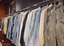 A neatly organized row of jackets hanging on a clothing rack, featuring a variety of denim, tan, and neutral colors. The wooden wall and shelves add a warm touch to the setting, with a few pairs of sandals placed below.