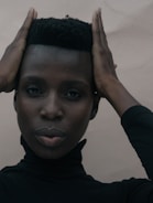 A person with a calm expression holds their hands on the sides of their head, wearing a black turtleneck against a neutral background.
