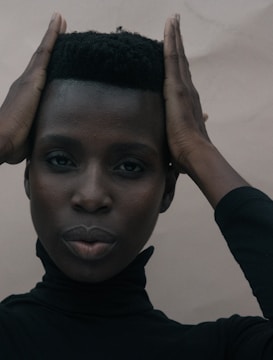 A person with a calm expression holds their hands on the sides of their head, wearing a black turtleneck against a neutral background.