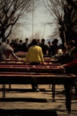 A group of people engaged in an outdoor activity involving multiple tables set up in parallel rows. The central figure wears a bright yellow jacket, standing with outstretched arms as other individuals observe or participate. Surrounding trees are bare, suggesting a season transition, and there's a sense of busyness and focus among the participants.