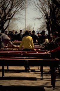 A group of people engaged in an outdoor activity involving multiple tables set up in parallel rows. The central figure wears a bright yellow jacket, standing with outstretched arms as other individuals observe or participate. Surrounding trees are bare, suggesting a season transition, and there's a sense of busyness and focus among the participants.