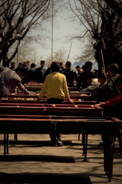 A group of people engaged in an outdoor activity involving multiple tables set up in parallel rows. The central figure wears a bright yellow jacket, standing with outstretched arms as other individuals observe or participate. Surrounding trees are bare, suggesting a season transition, and there's a sense of busyness and focus among the participants.