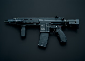A modern black assault rifle with a flashlight attachment is displayed against a dark background. The weapon has a short barrel and a tactical design with various components including a magazine, grip, and stock.