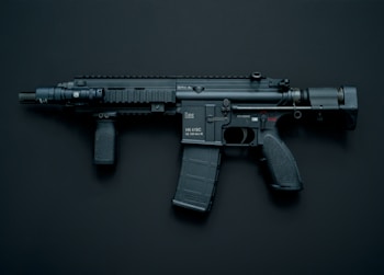 A modern black assault rifle with a flashlight attachment is displayed against a dark background. The weapon has a short barrel and a tactical design with various components including a magazine, grip, and stock.