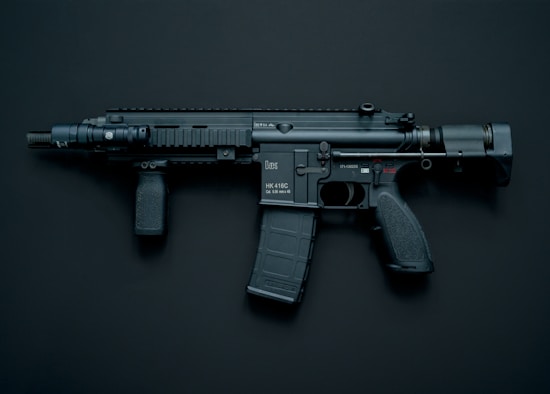A modern black assault rifle with a flashlight attachment is displayed against a dark background. The weapon has a short barrel and a tactical design with various components including a magazine, grip, and stock.