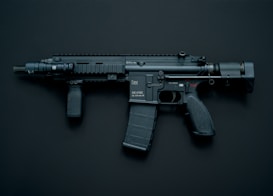 A modern black assault rifle with a flashlight attachment is displayed against a dark background. The weapon has a short barrel and a tactical design with various components including a magazine, grip, and stock.
