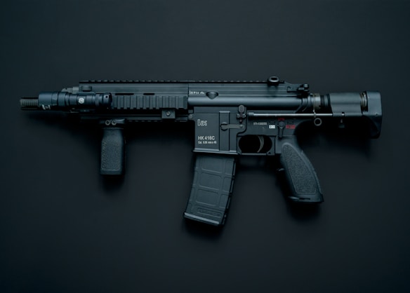 A modern black assault rifle with a flashlight attachment is displayed against a dark background. The weapon has a short barrel and a tactical design with various components including a magazine, grip, and stock.