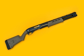 A tactical shotgun with a black metal barrel and olive green synthetic stock and fore-end is displayed diagonally on a bright yellow background. The shotgun features a pump-action design with a sleek, modern appearance.