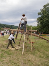 Two individuals engaged in a traditional wood-cutting activity using a large saw. One is standing on a wooden structure, while the other is on the ground. The scene takes place outdoors in a grassy area with a small audience watching in the background. The people are dressed in period-style clothing and a rural landscape with trees and hills is visible.