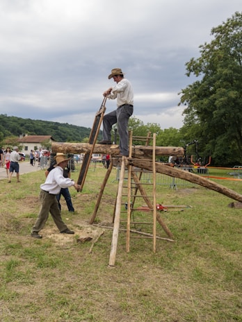 Two individuals engaged in a traditional wood-cutting activity using a large saw. One is standing on a wooden structure, while the other is on the ground. The scene takes place outdoors in a grassy area with a small audience watching in the background. The people are dressed in period-style clothing and a rural landscape with trees and hills is visible.