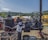 A person wearing historical clothing operates or maintains a large, antique steam engine outdoors at a fair or machinery exhibition. In the background, there are tents, people, and inflatable structures. The setting appears to be rural with hills and trees in the distance.