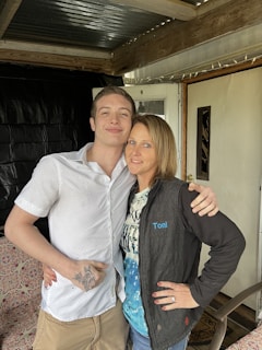 A young man and a woman stand close together, smiling, under a covered porch with wooden beams. The young man is wearing a white, short-sleeved shirt and has a tattoo on his hand. The woman is wearing a black jacket with the name 'Toni' embroidered on it over a patterned blue and white shirt. They are both embracing each other and appear relaxed and happy.