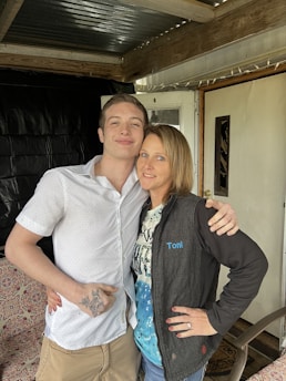 A young man and a woman stand close together, smiling, under a covered porch with wooden beams. The young man is wearing a white, short-sleeved shirt and has a tattoo on his hand. The woman is wearing a black jacket with the name 'Toni' embroidered on it over a patterned blue and white shirt. They are both embracing each other and appear relaxed and happy.