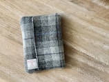 A plaid fabric item, likely a wallet or small pouch, rests on a wooden surface. The fabric features a pattern of grey, black, and blue squares with an attached label reading 'Harris Tweed'.