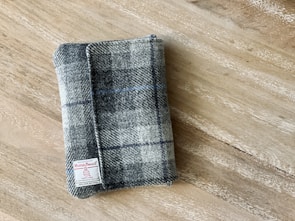 A plaid fabric item, likely a wallet or small pouch, rests on a wooden surface. The fabric features a pattern of grey, black, and blue squares with an attached label reading 'Harris Tweed'.